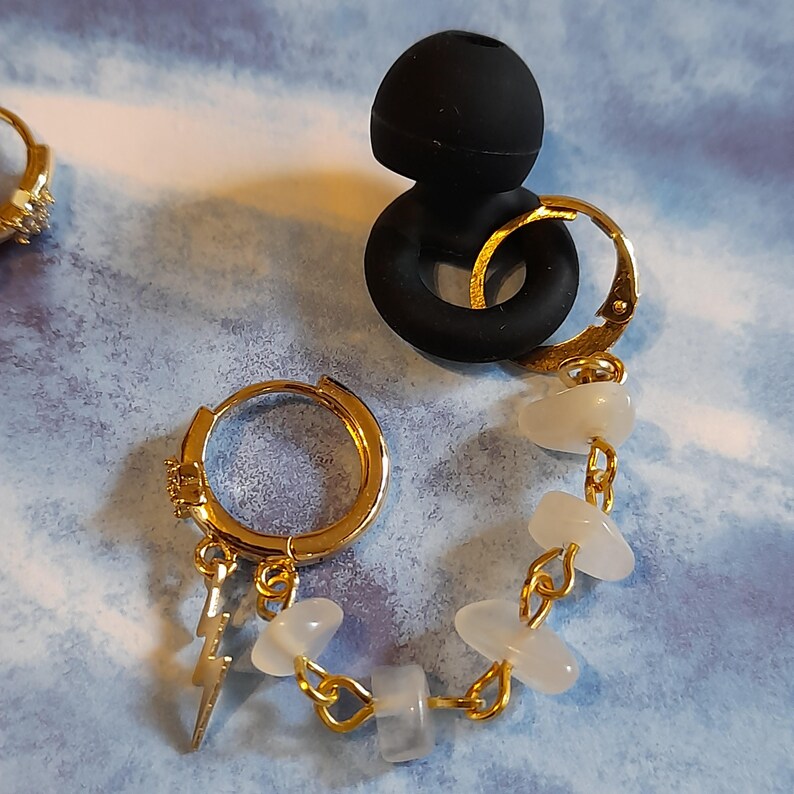 May include: A gold hoop earring featuring a dangling lightning bolt charm and a chain of small, translucent white stone beads. The earring is connected to a black silicone earbud tip. A second gold hoop earring with small clear stones is visible.