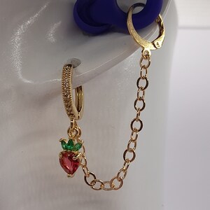 May include: Gold hoop earrings with a dangling red strawberry charm with green leaves and clear rhinestones. The earrings are connected by a gold chain.