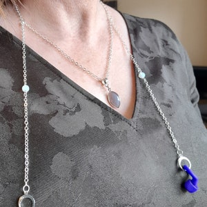 May include: A silver chain necklace with a gray stone pendant and a second chain with a blue earbud holder. The necklace is worn by a person with a floral patterned shirt.