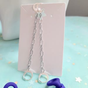 May include: Silver-toned dangle earrings. Each earring features a chain of elongated oval links, suspending a small, open circle at the bottom. The earrings are displayed on a white card against a light blue background.