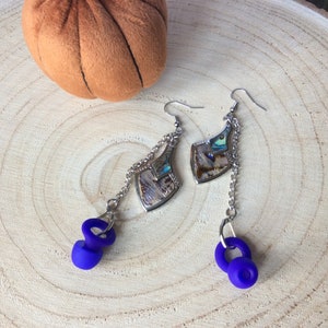 May include: Silver earrings featuring iridescent abalone shell details and hanging blue spiral elements. The earrings are suspended from chains. They are presented on a wooden surface, with a small orange pumpkin in the background.