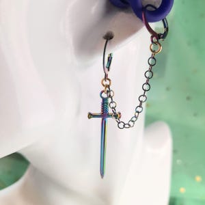 May include: A single earring featuring a small, iridescent sword pendant. The sword is connected to a chain that attaches to a hoop earring. The earring has a rainbow metallic finish, with colors shifting from blue to green and purple.