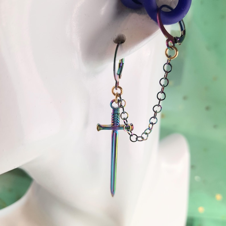 May include: A single earring featuring a small, iridescent sword pendant. The sword is connected to a chain that attaches to a hoop earring. The earring has a rainbow metallic finish, with colors shifting from blue to green and purple.