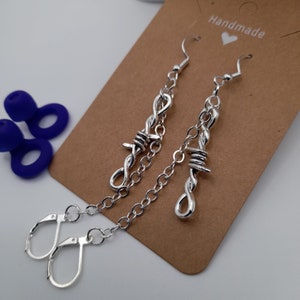 May include: A pair of silver earrings featuring a barbed wire design. The earrings hang from a chain and have a hook fastening. The earrings are handmade.