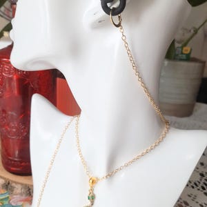 May include: A gold-toned chain earring connector that links a black ear plug to a gold snake pendant necklace. The snake charm features small green eyes and a textured body. The delicate metal chain extends from the ear to the necklace pendant.
