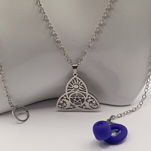 May include: A silver necklace with a pendant featuring a triple moon, pentacle, and sun design. The necklace is on a white surface with a pair of blue silicone ear plugs.