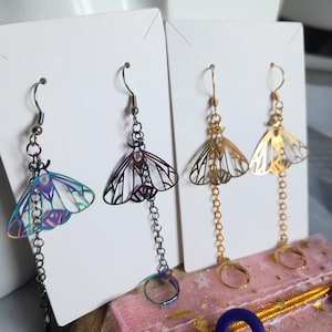 May include: Earrings featuring a moth design. One pair has a rainbow iridescent finish, another is black, and the last two are gold. Each earring has a chain and a hoop at the bottom. The earrings are displayed on white cards.