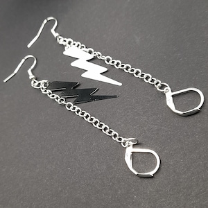May include: A pair of silver earrings with black and white lightning bolt charms hanging from silver chains. The earrings have a hook closure.