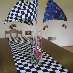 May include: A black and white chequered table runner with a vase filled with toy race cars and chequered flags. The flags are black and white and blue with monster truck designs.