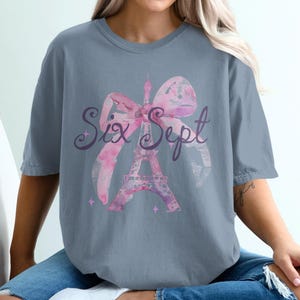 May include: A slate blue t-shirt featuring a watercolor design of the Eiffel Tower and a pink bow. The words "Six Sept" are written in a cursive font across the design. The shirt is a relaxed fit.