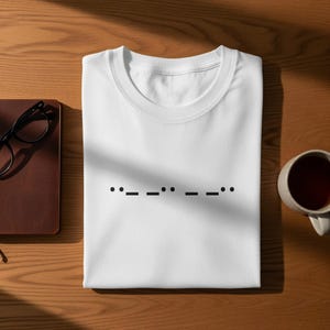 May include: White t-shirt with a black Morse code design. The shirt is folded neatly on a wooden surface, with a leather-bound notebook, eyeglasses, and a mug of tea nearby. The Morse code spells out a word.