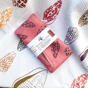 May include: A reversible hand towel with a coral-colored side featuring a brown seashell pattern. The towel is folded and wrapped with a white band. The background is a white towel with various seashell designs in brown, orange, and red.