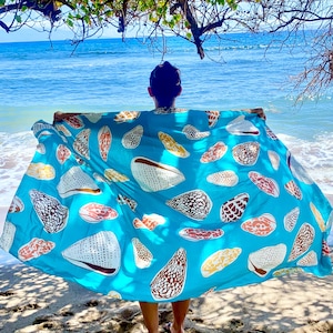 May include: A turquoise beach towel featuring a seashell pattern in shades of white, brown, and yellow. The towel is held up against a backdrop of the ocean and a sandy beach, creating a summery vibe.