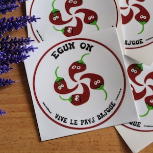 May include: Five red chili peppers with faces form a five-pointed star. The star is surrounded by a red circle with the text "EGUN ON" in white. Below the circle is the text "VIVE LE PAYS BASQUE" in white.