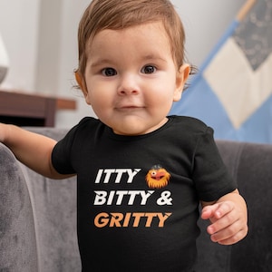 May include: A black baby onesie with the text "itty bitty & gritty" in white and orange. A cartoon orange mascot with a black eye patch is next to the text.