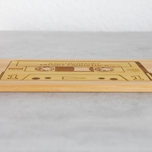 Puede incluir: Una tabla de cortar de madera con un dise&ntilde;o de cinta de casete. La cinta est&aacute; etiquetada como "Bastelkinder Tatort Fr&uuml;hst&uuml;ck".