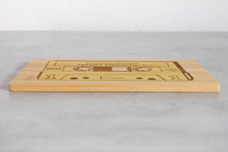 Puede incluir: Una tabla de cortar de madera con un dise&ntilde;o de cinta de casete. La cinta est&aacute; etiquetada como "Bastelkinder Tatort Fr&uuml;hst&uuml;ck".