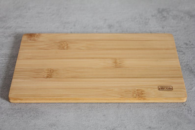 Puede incluir: Una tabla de cortar rectangular de bamb&uacute; marr&oacute;n claro con las palabras "Live Your Dreams" impresas en una peque&ntilde;a etiqueta rectangular en la esquina inferior derecha.