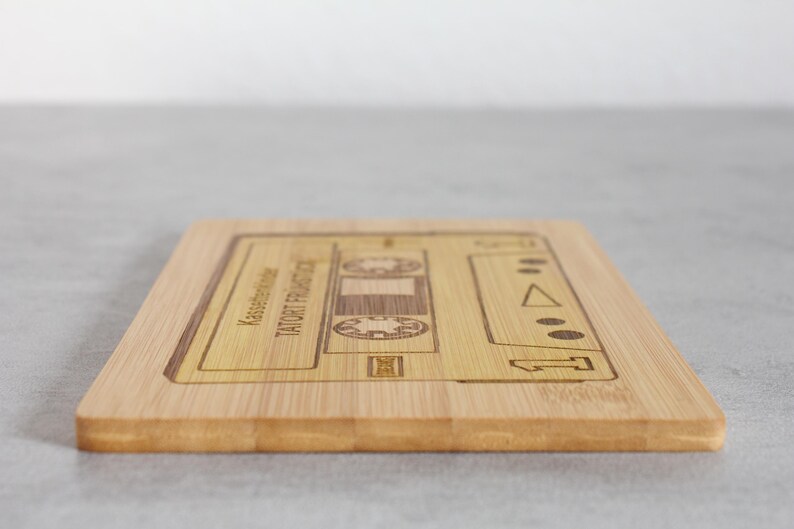 Puede incluir: Una tabla de cortar de madera con un dise&ntilde;o de cinta de casete. El texto "TAUGHT FROM THE HEART" y "Kassenbon" est&aacute; grabado en la tabla.