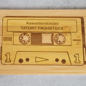 Puede incluir: Una tabla de cortar de madera con un dise&ntilde;o de cinta de casete. La cinta est&aacute; etiquetada como "Kassettenkinder TATOR FR&Uuml;HST&Uuml;CK" y tiene las palabras "DREAMS" y "stereo" impresas en ella.