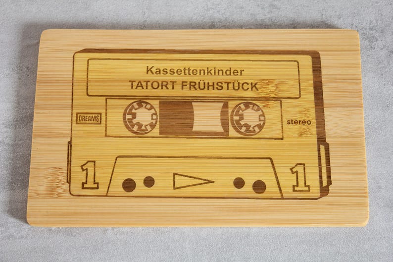 Puede incluir: Una tabla de cortar de madera con un dise&ntilde;o de cinta de casete. La cinta est&aacute; etiquetada como "Kassettenkinder TATOR FR&Uuml;HST&Uuml;CK" y tiene las palabras "DREAMS" y "stereo" impresas en ella.