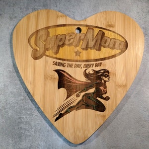 Puede incluir: Una tabla de cortar de bambú en forma de corazón con un diseño grabado con láser. El diseño presenta a una superheroína volando con el texto "Super Mom" y "Saving the day, every day".