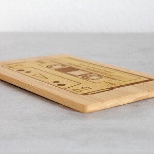 Puede incluir: Una tabla de cortar de madera con una imagen de una cinta de casete grabada con l&aacute;ser. La tabla es de color marr&oacute;n claro y tiene una superficie lisa.