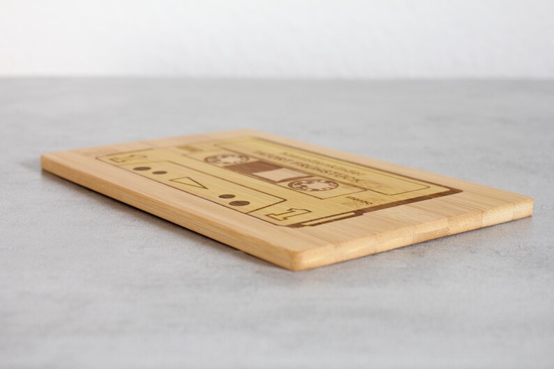 Puede incluir: Una tabla de cortar de madera con una imagen de una cinta de casete grabada con l&aacute;ser. La tabla es de color marr&oacute;n claro y tiene una superficie lisa.