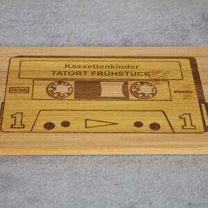 Puede incluir: Una tabla de cortar de madera con un dise&ntilde;o de cinta de casete. La cinta est&aacute; etiquetada como "Kassettenkinder TATOR FR&Uuml;HST&Uuml;CK" y tiene las palabras "DR.DREAMS" y "stereo" impresas en ella.