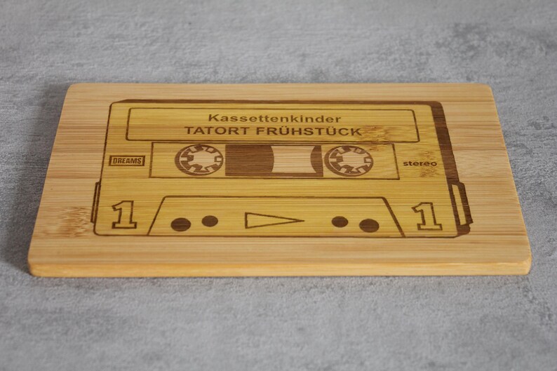 Puede incluir: Una tabla de cortar de madera con un dise&ntilde;o de cinta de casete. La cinta est&aacute; etiquetada como "Kassettenkinder TATOR FR&Uuml;HST&Uuml;CK" y tiene las palabras "DR.DREAMS" y "stereo" impresas en ella.