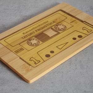Puede incluir: Una tabla de cortar de madera con forma de cinta de casete con el texto "Kassettenkinder TATORT FR&Uuml;HST&Uuml;CK" grabado en ella.
