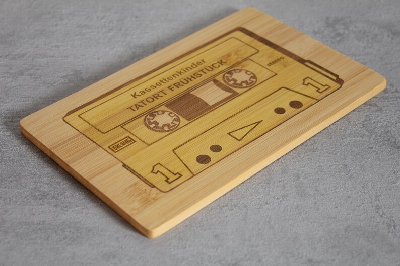 Puede incluir: Una tabla de cortar de madera con forma de cinta de casete con el texto "Kassettenkinder TATORT FR&Uuml;HST&Uuml;CK" grabado en ella.