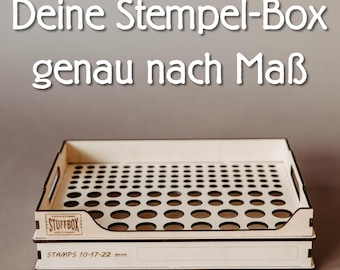 May include: A light-colored wooden storage tray featuring a grid of circular cutouts of varying sizes. The front panel displays the text STUFFBOX and STAMPS 10-17-22 mm. The top of the image includes the German text Deine Stempel-Box genau nach Maß.