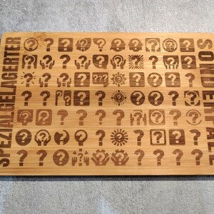 Puede incluir: Tabla de cortar rectangular de bambú con diseños de signos de interrogación grabados. Las palabras "SPEZIAL GELAGERTER" y "SONDERFALL" están grabadas verticalmente. La tabla es de color marrón claro.