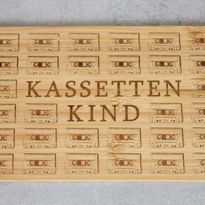 Puede incluir: Una tabla de cortar de madera con un diseño retro que presenta múltiples ilustraciones de cintas de cassette. La tabla está grabada con el texto "KASSETTEN KIND".