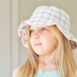 May include: A young girl with long blonde hair wearing a white and blue gingham sun hat with a floral print lining. She is wearing a turquoise tank top with a white pattern.