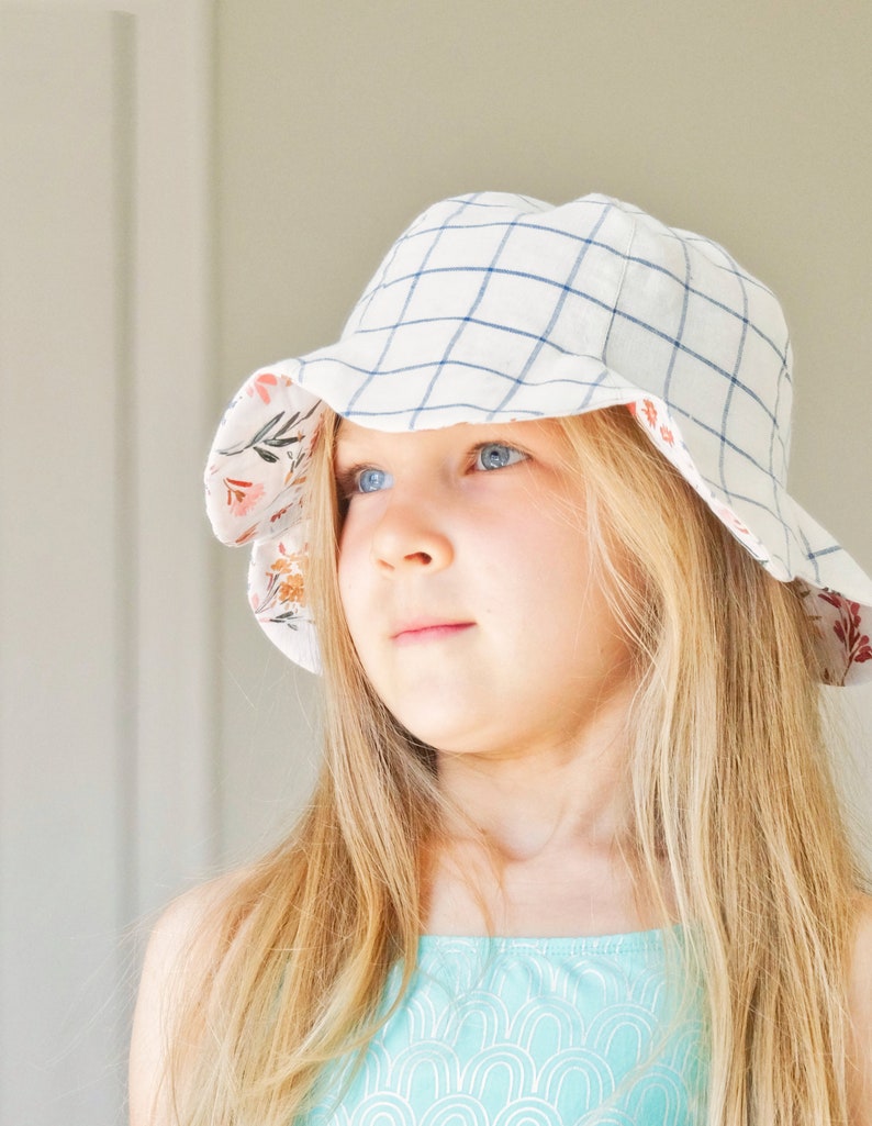 May include: A young girl with long blonde hair wearing a white and blue gingham sun hat with a floral print lining. She is wearing a turquoise tank top with a white pattern.