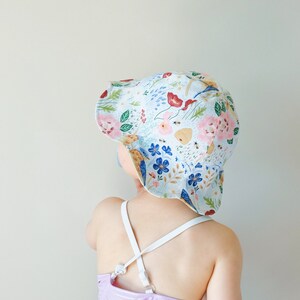 May include: A child wearing a white sun hat with a floral pattern and a purple swimsuit. The hat has a wide brim and is decorated with flowers, bees, and birds.