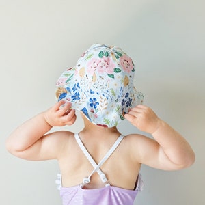 May include: A child wearing a white sun hat with a floral pattern and a lavender swimsuit. The hat has a wide brim and is pulled down over the child's eyes.
