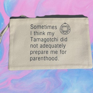 Op de afbeelding: Een beige canvas draagtas met een zwarte rits en een zwarte handvat. De tas heeft een zwart-witte afbeelding van een Tamagotchi en de tekst "Sometimes I think my Tamagotchi did not adequately prepare me for parenthood."