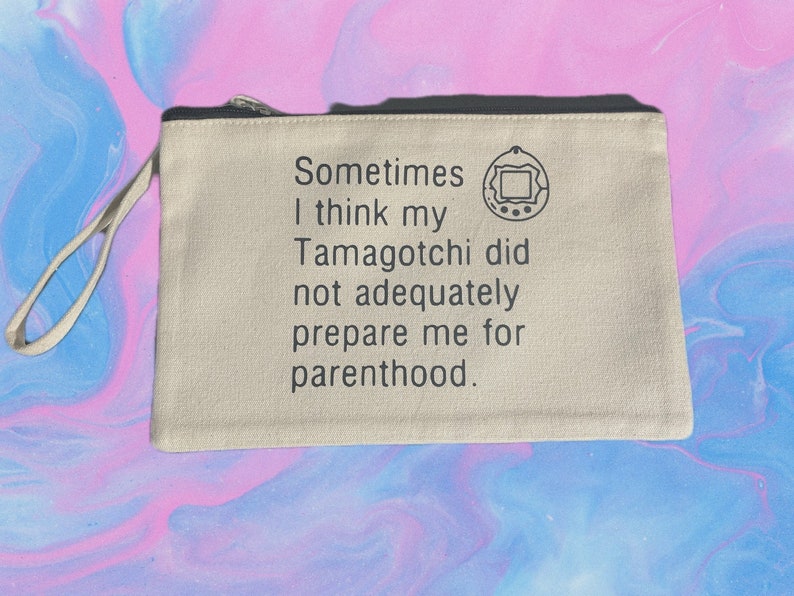 Op de afbeelding: Een beige canvas draagtas met een zwarte rits en een zwarte handvat. De tas heeft een zwart-witte afbeelding van een Tamagotchi en de tekst "Sometimes I think my Tamagotchi did not adequately prepare me for parenthood."
