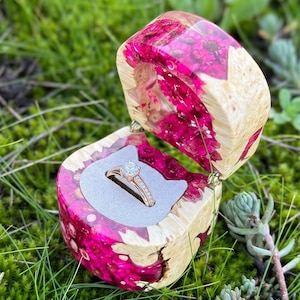 May include: A ring box crafted from light-coloured wood and vibrant fuchsia resin, featuring a hinged lid and a grey interior insert. A metallic ring with a central stone and small accent stones is positioned inside the open ring box.