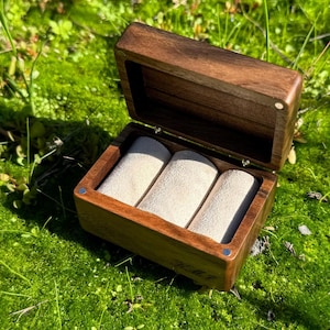Puede incluir: Caja de reloj de madera abierta con tres soportes acolchados beige. La caja es de madera marrón oscuro y se encuentra sobre un lecho de musgo verde. La caja está abierta, revelando el interior. La caja tiene las iniciales "M.M.J" grabadas.