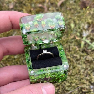 May include: A square ring box made of clear resin embedded with green moss and small plants, featuring a hinged lid and a black velvet interior insert holding a thin metal ring with a single small clear stone set in the center.