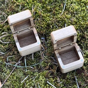 May include: Two small, open wooden boxes with hinged lids. The boxes are square with rounded edges and a light wood finish. The interiors are a darker wood. The boxes are set on a bed of green moss.