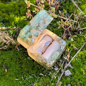 Puede incluir: Una caja de anillos decorativa abierta con un anillo de oro engastado con una piedra preciosa azul, que descansa sobre un cojín beige. La caja está hecha de un material de color claro con un diseño verde y azul veteado. La caja está en musgo verde.