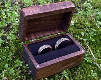May include: Two rings with a dark wood and metallic inlay design are nestled inside a rectangular dark brown wooden box. The box features a hinged lid and a black velvet interior lining, providing a secure space for the two circular bands.
