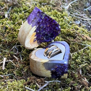 Può includere: Scatola portaanelli in legno a forma di cuore con parte superiore in resina viola, contenente due anelli d'argento. La scatola è aperta, rivelando gli anelli appoggiati su una superficie grigia. La scatola è su un letto di muschio verde.
