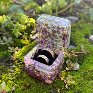 May include: A square resin ring box featuring embedded dried flowers in shades of purple, pink, and white. The box is open, revealing a black velvet interior holding two metallic rings with a dark band and a thin gold-colored center stripe.