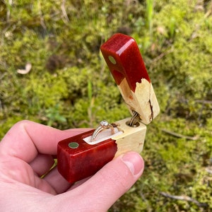 May include: A handcrafted wooden ring box with a red resin top and a natural wood base. The box is open, revealing a gold ring with a diamond center stone. The box is held against a background of green moss.